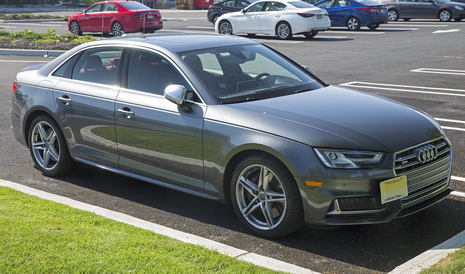 Dark gray Audi S4 sedan parked in a sunny parking lot next to a strip of grass, seen from the front three-quarter angle.