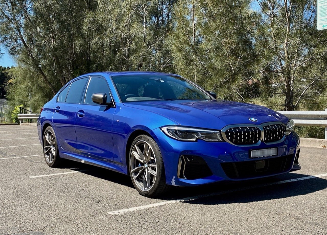 Blue BMW M340i sedan parked in an empty parking lot, seen from the front three-quarter angle with tall trees behind it.