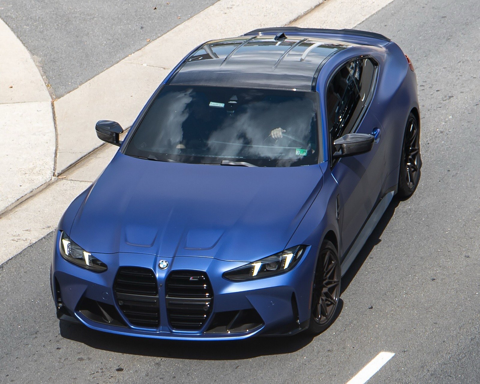 A matte blue BMW M4 Competition coupe, seen from above, drives along a street beside a curb and sidewalk.