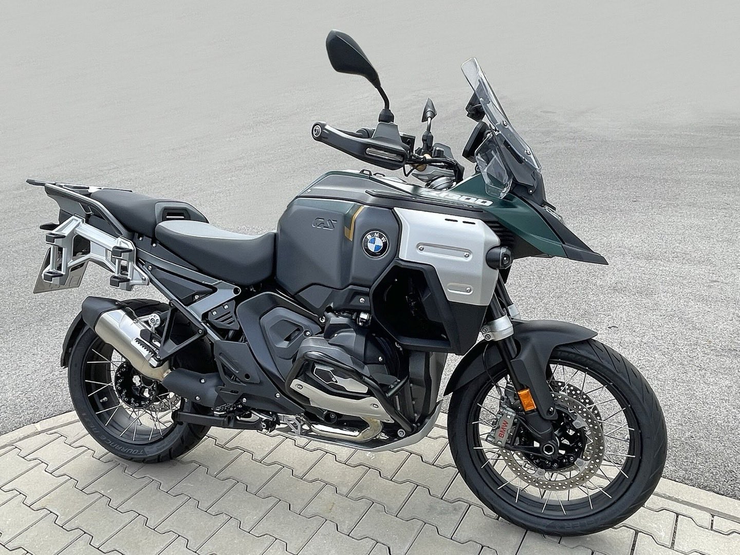 Grey and green BMW R 1300 GS Adventure motorcycle with spoked wheels and luggage rack, parked on paving stones.