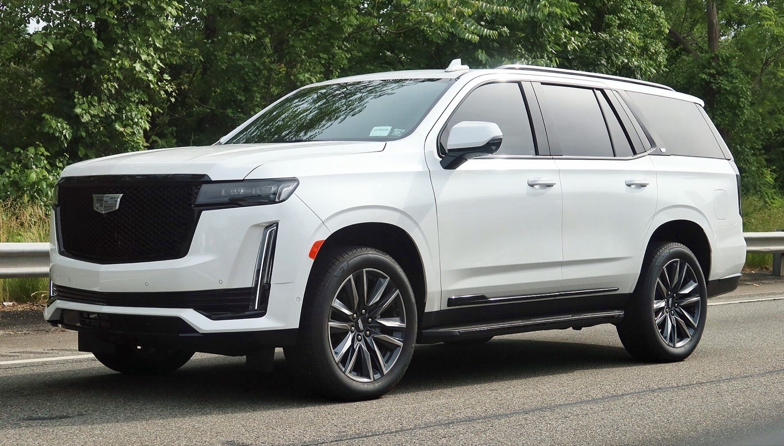 White 2021 Cadillac Escalade on a road lined with a guardrail and green trees, seen from the front three-quarter angle.