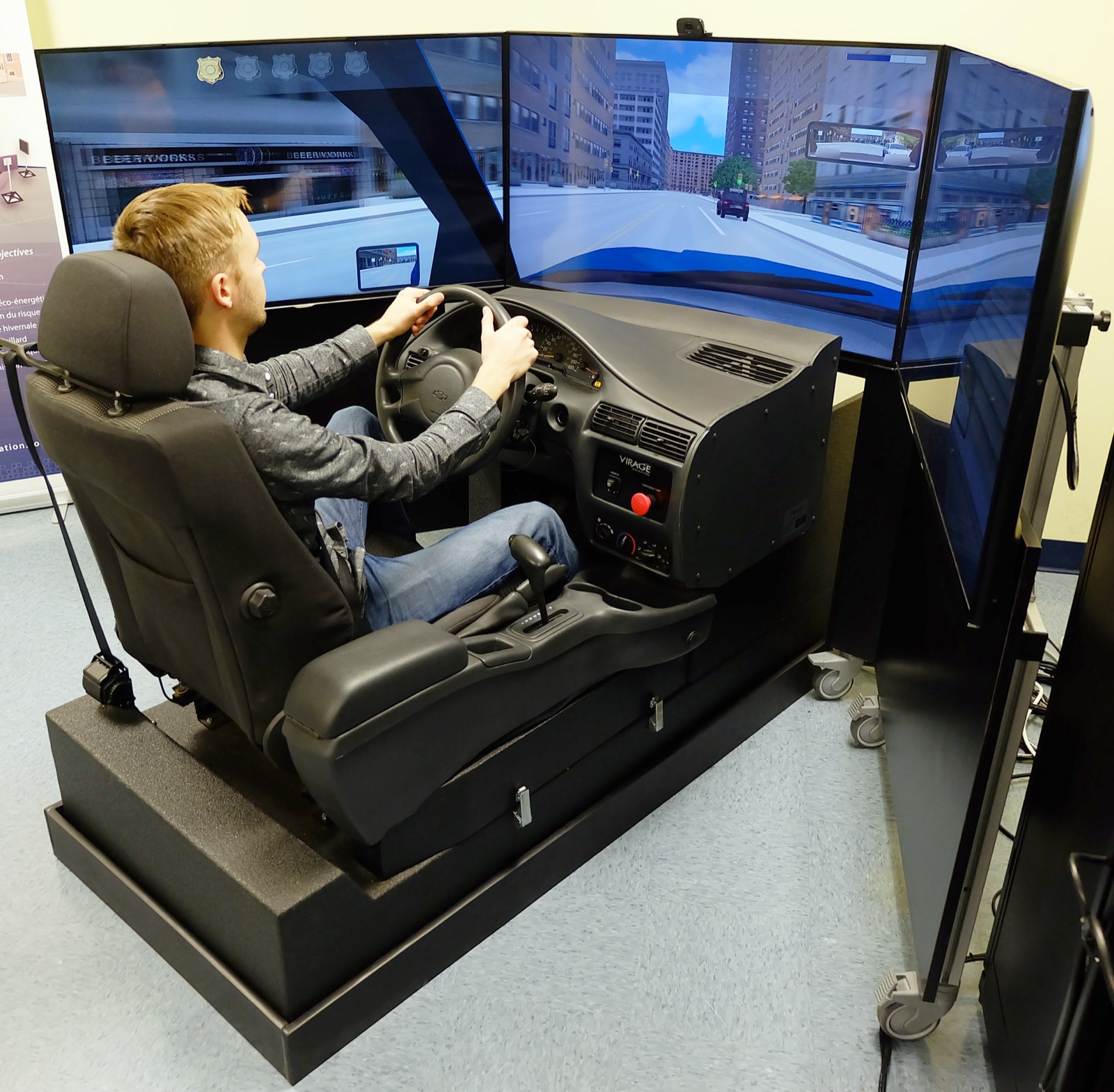 A young man sits at the wheel of a driver-training simulator, facing three curved screens that show a city street from the driver's seat.