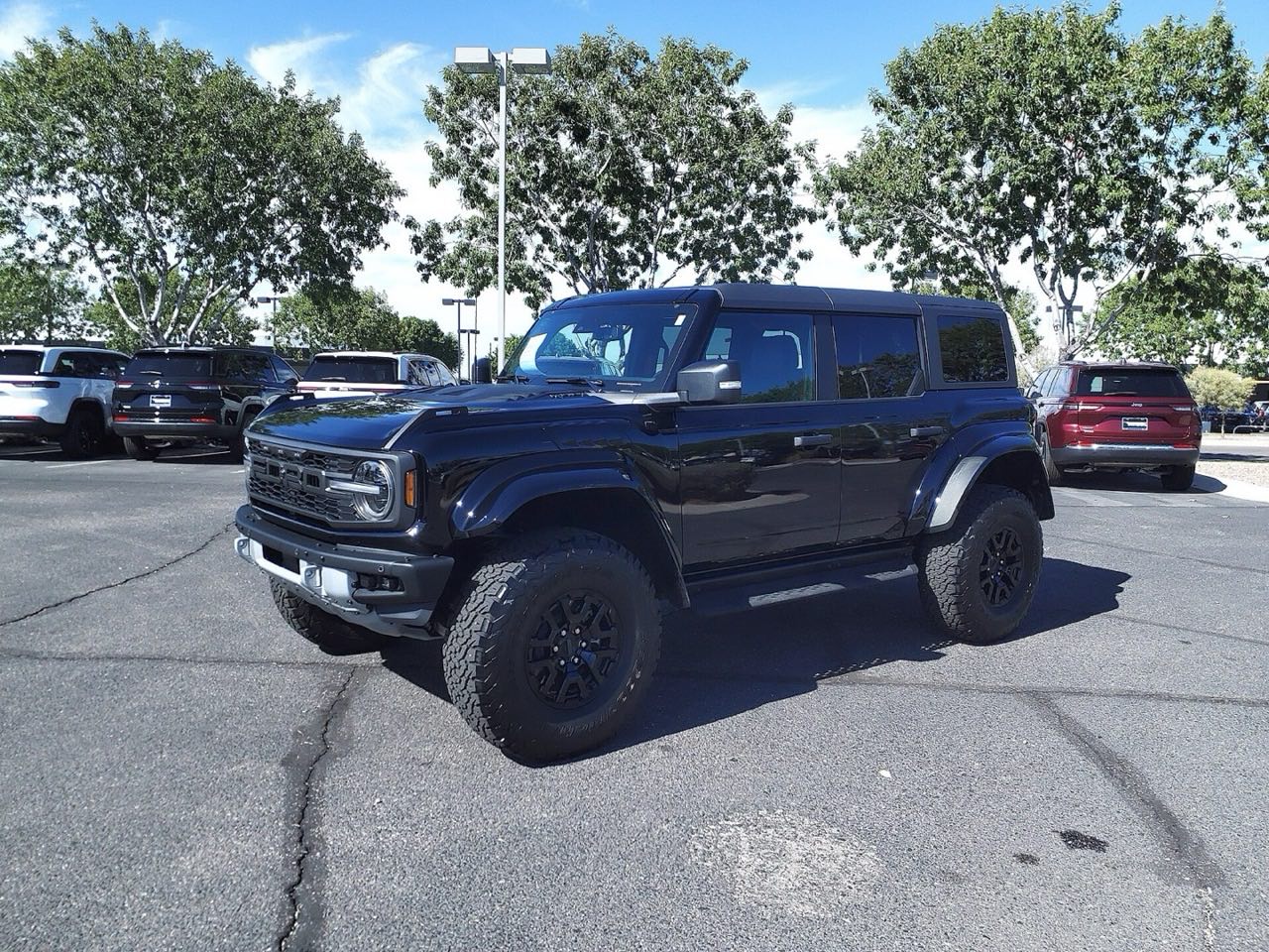 Black 2024 Ford Bronco Raptor parked in a sunny outdoor parking lot with trees behind it, seen from the front three-quarter angle.