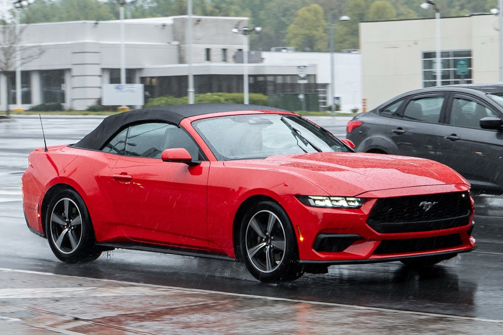 A red Ford Mustang convertible with its black soft top raised drives along a wet road in the rain, past a parked car and low commercial buildings.