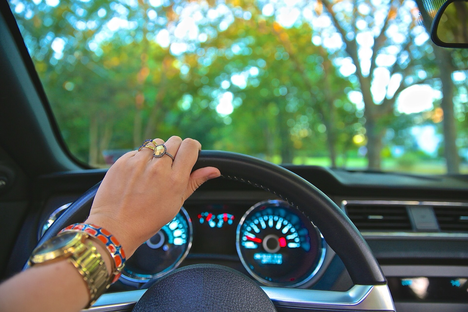 A driver's hand, wearing a gold watch and bracelets, rests on top of the steering wheel above a lit instrument cluster, with a tree-lined road visible through the windshield.