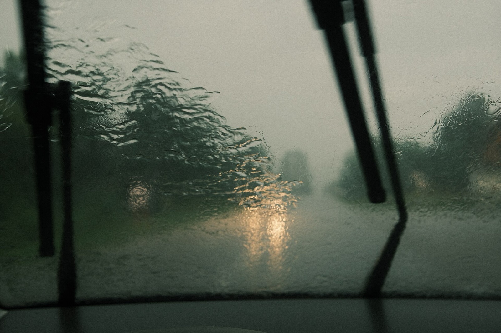 Heavy rain on a car windshield, the wipers mid-sweep and the road ahead blurred by water and headlight glare.