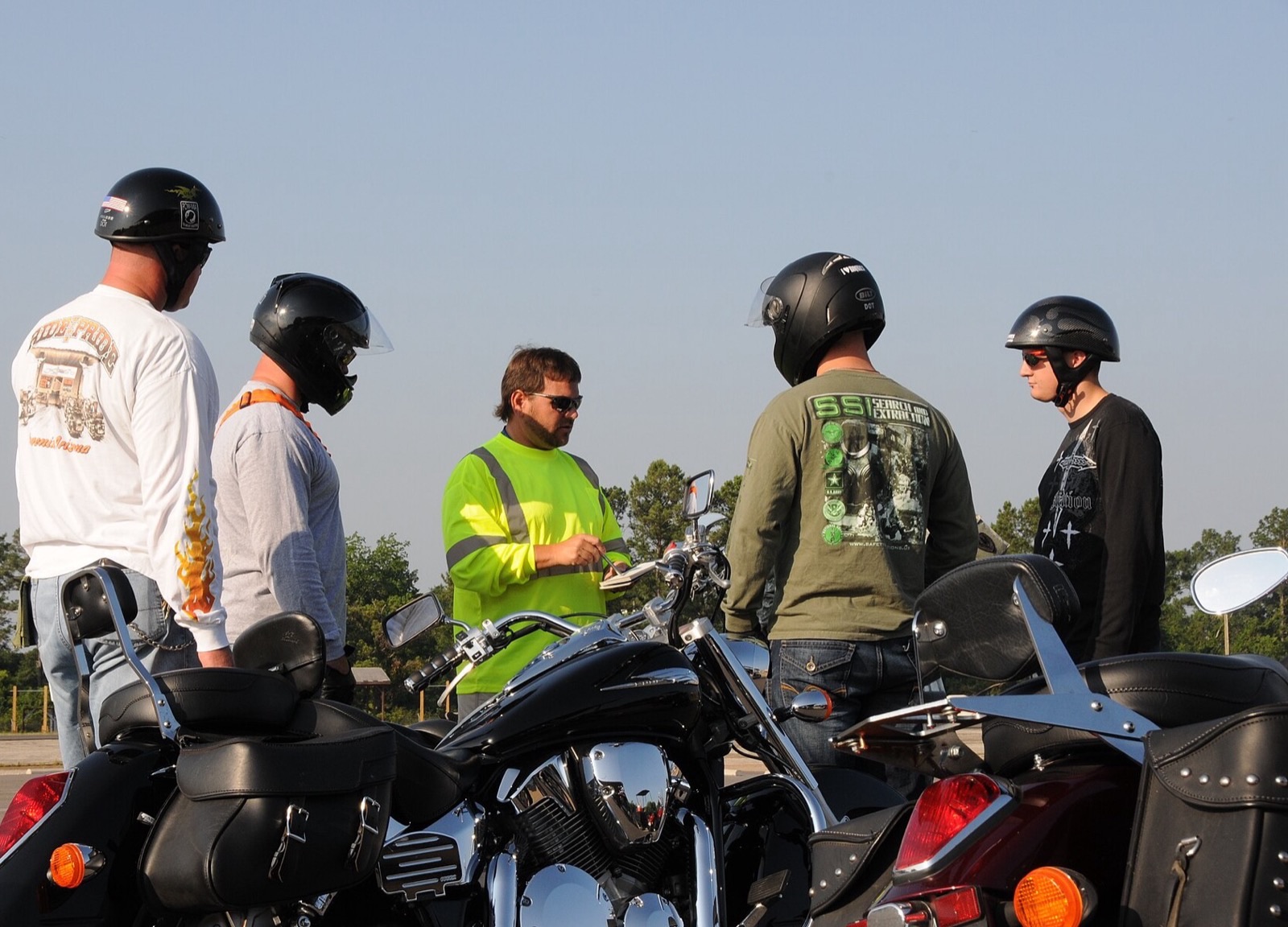 A coach in a high-visibility yellow shirt reviews notes with four helmeted riders standing beside parked touring motorcycles on an open practice lot.