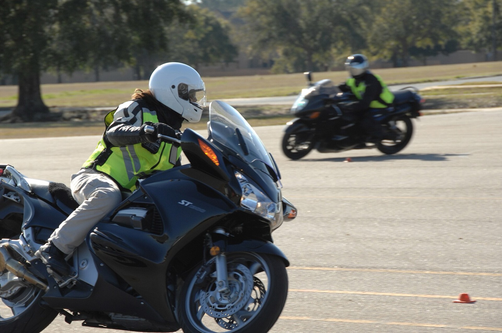 Two motorcycle riders in full-face helmets and high-visibility vests practicing on a paved training range marked with small cones.
