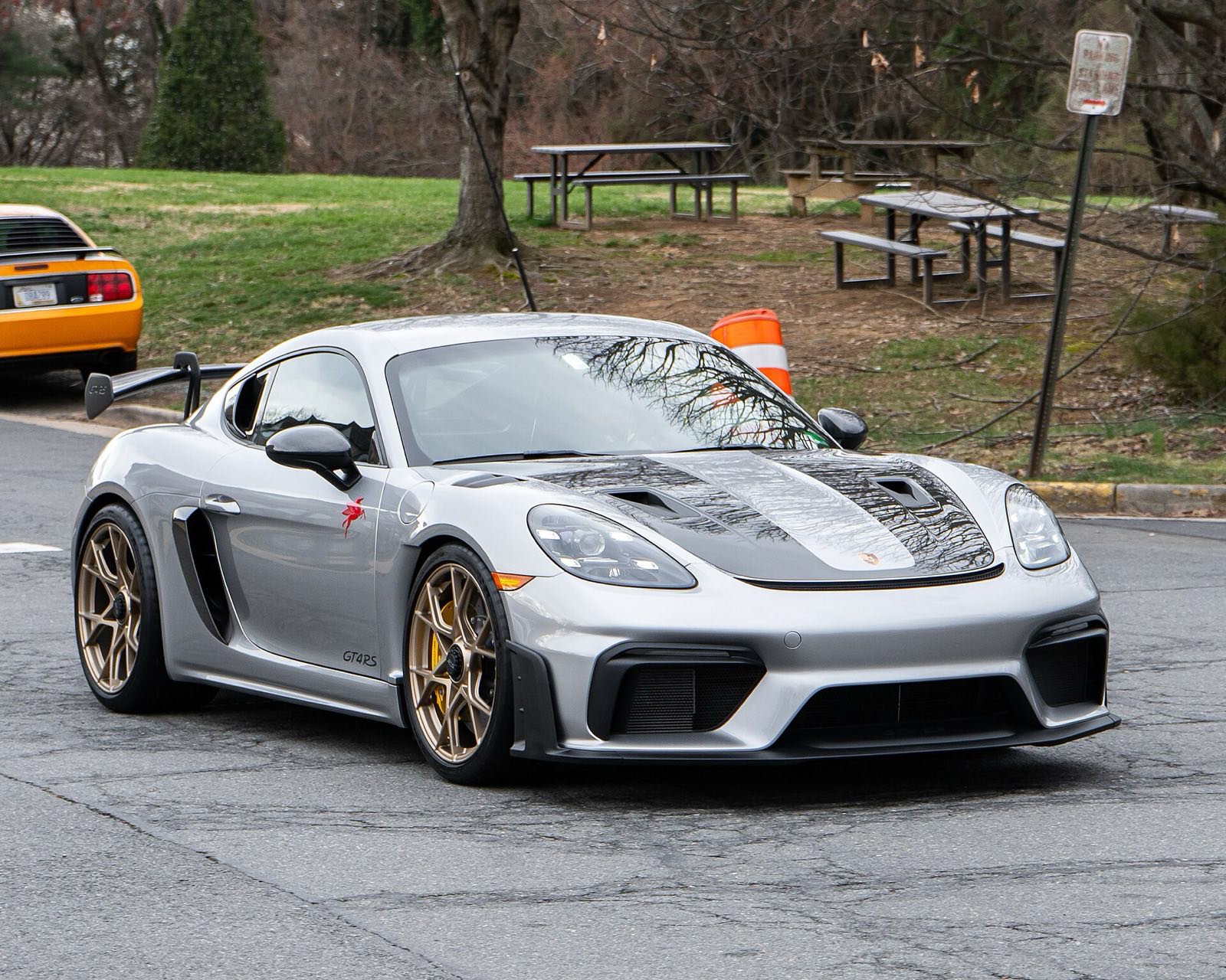 A silver Porsche 718 Cayman GT4 RS with bronze wheels and a large rear wing drives along a park road past bare trees, picnic tables and an orange traffic barrel.
