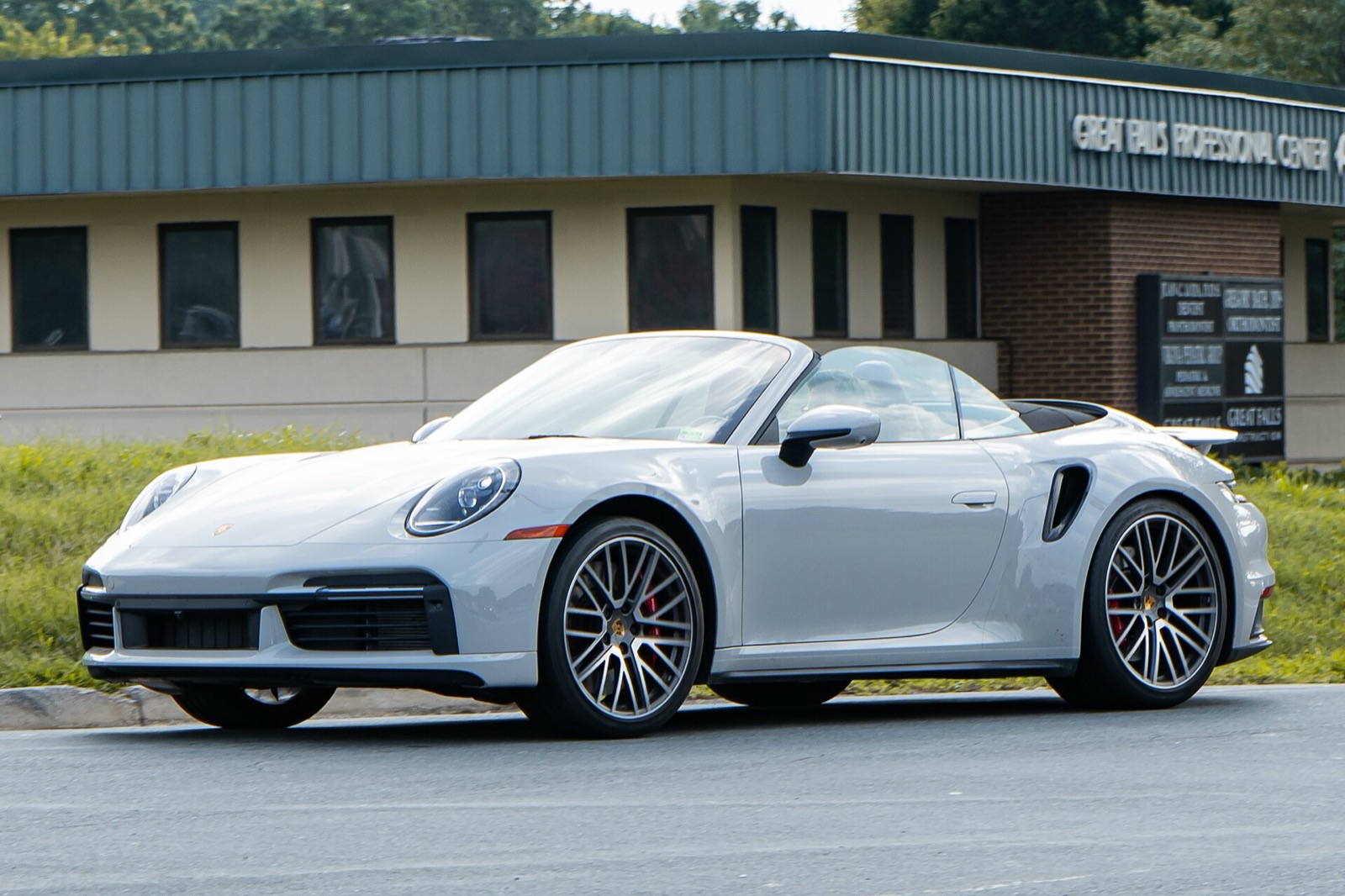 A pale grey Porsche 911 Turbo Cabriolet with its roof down on a suburban road, in front of a grassy verge and a low office building.