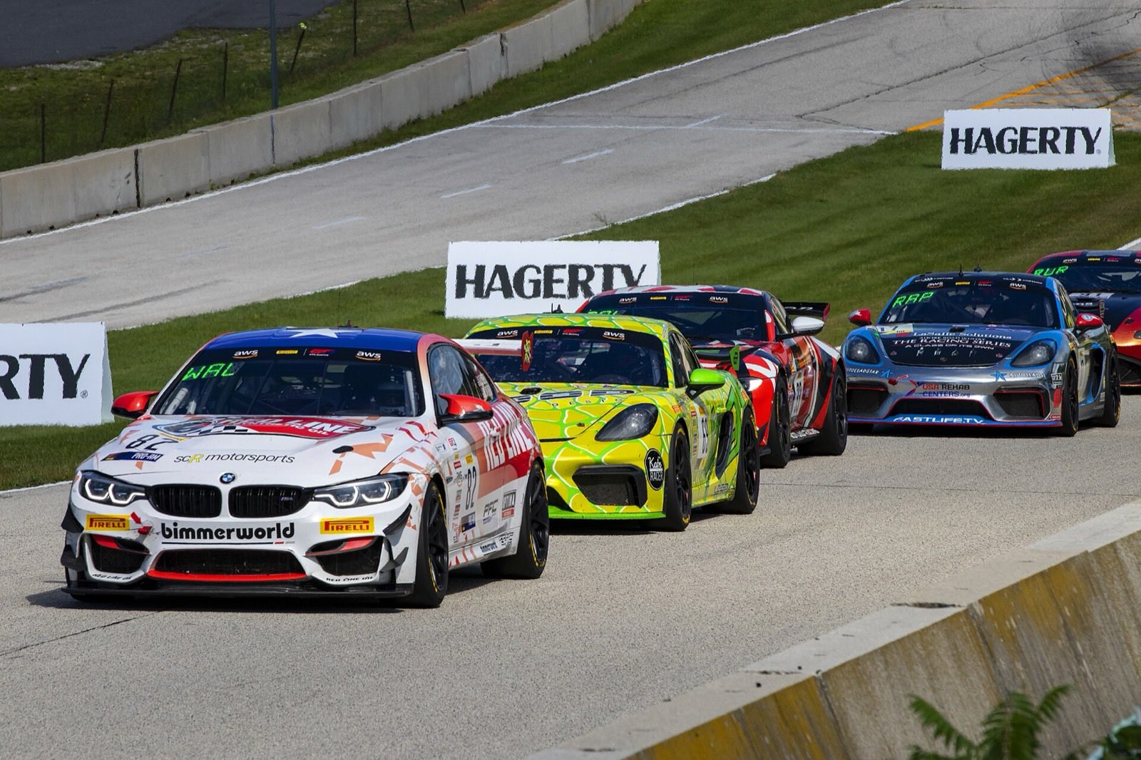 A BMW M4 GT4 race car leads several Porsche 718 Cayman GT4 race cars through a corner at the Road America road course.
