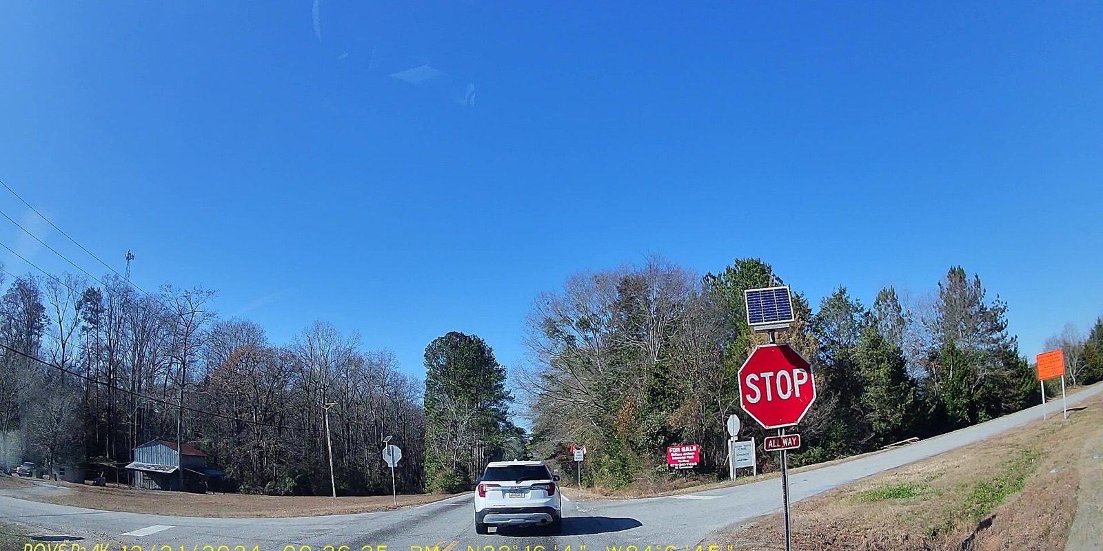 A driver’s-eye view of a two-lane road meeting a stop sign at a rural intersection, with a white SUV waiting ahead.